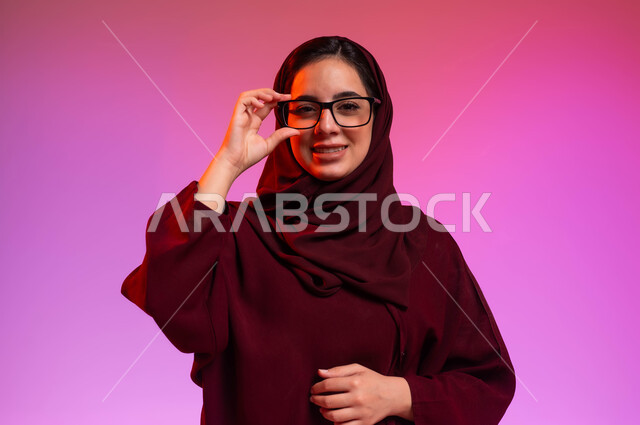المحافظة على صحة العينين، ارتداء النظارات الخاصة بضعف النظر، بورتريه مقرب لسيدة عربية خليجية سعودية مبتسمة محجبة ترتدي العباءة تضع النظاره الطبيه وتنظر إلى الكاميرا بإيماءات السرور، مفهوم تحسين الرؤية ومحاولة تعديل البصر، خلفية ملونة