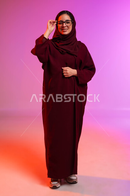 مفهوم تحسين الرؤية ومحاولة تعديل البصر، المحافظة على صحة العينين، ارتداء النظارات الخاصة بضعف النظر، بورتريه لسيدة عربية خليجية سعودية مبتسمة محجبة ترتدي العباءة تضع النظاره الطبيه وتنظر إلى الكاميرا بإيماءات السرور، صورة الجسم بالطول الكامل، خلفية ملونة