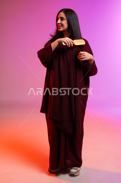 Care and attention to beauty and elegance, expressions and gestures that indicate happiness and pleasure, maintaining healthy and elegant hair, a portrait of a smiling Saudi Arabian Gulf woman styling and combing her hair using a brush (comb), full body portrait, colored background