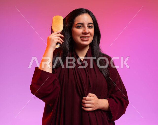 النظر إلى الكاميرا بإيماءات تدل على السعادة والسرور، المحافظة على شعر صحي وأنيق، العناية والاهتمام بالجمال والأناقة والمظهر الخارجي، بورتريه مقرب لشابة عربية خليجية سعودية مبتسمة تقوم بتصفيف وتمشيط شعرها باستخدام الفرشاة ( المشط )، خلفية ملونة