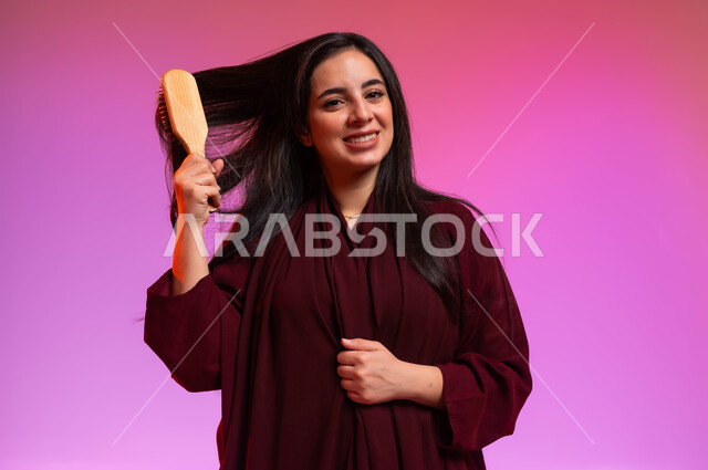 النظر إلى الكاميرا بإيماءات تدل على السعادة والسرور، المحافظة على شعر صحي وأنيق، العناية والاهتمام بالجمال والأناقة والمظهر الخارجي، بورتريه مقرب لشابة عربية خليجية سعودية مبتسمة تقوم بتصفيف وتمشيط شعرها باستخدام الفرشاة ( المشط )، خلفية ملونة