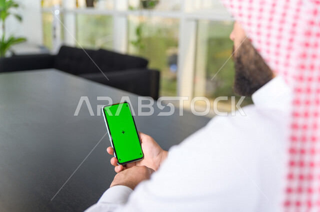 رجل أعمال سعودي خليجي يستخدام هاتفه المحمول بخلفية خضراء كروما ، تقنية و حاسب ، استخدام تقنية السمارت على الهاتف المحمول ليتحكم بالتلفاز