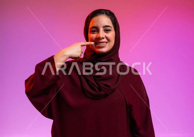إبتسامة هوليود باستخدام قشور فينير من الخزف، بورتريه مقرب لشابة عربية خليجية سعودية مبتسمة ترتدي العباءة والحجاب تنظر إلى الكاميرا وتشير بإصبع السبابة على أسنانها، العناية والاهتمام بالصحة الفموية، خلفية ملونة.
