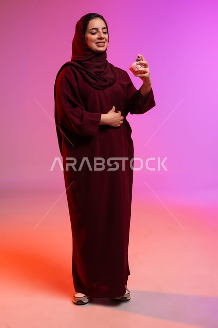 التطيب والتعطر بالعود والزيوت العطرية, التسويق لمنتجات العطور والمستحضرات النسائية, مفهوم الأناقة والاهتمام بالمظهر الخارجي, بورتريه لسيدة عربيه سعودية خليجية محجبة مبتسمة تقوم برش العطر بإيماءات الإعجاب بالروائح العطرة, صورة لكامل الجسم، خلفية ملونة