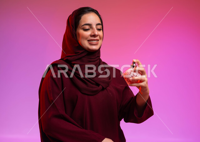 التسويق لمنتجات العطور والمستحضرات النسائية, مفهوم الأناقة والاهتمام بالمظهر الخارجي, بورتريه مقرب لسيدة عربيه سعودية خليجية محجبة مبتسمة تقوم برش العطر, ايماءات الإعجاب بالروائح العطرة, التطيب والتعطر بالعود والزيوت العطرية, خلفية ملونة