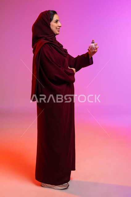 التطيب والتعطر بالعود والزيوت العطرية, التسويق لمنتجات العطور والمستحضرات النسائية, مفهوم الأناقة والاهتمام بالمظهر الخارجي, بورتريه من الجانب لسيدة عربيه سعودية خليجية محجبة مبتسمة تقوم برش العطر بإيماءات الإعجاب بالروائح العطرة, صورة لكامل الجسم، خلفية ملونة