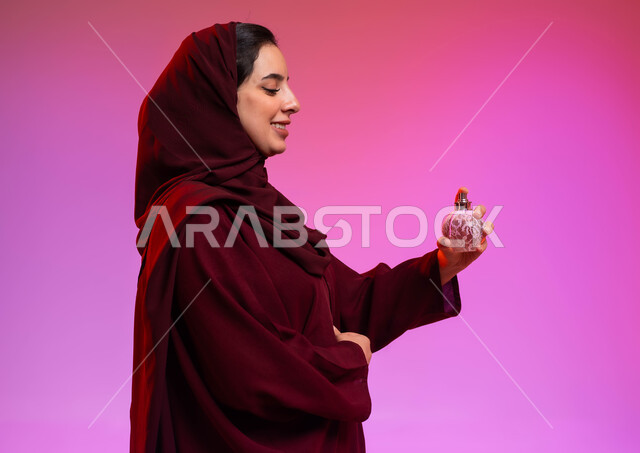 التطيب والتعطر بالعود والزيوت العطرية, التسويق لمنتجات العطور والمستحضرات النسائية, مفهوم الأناقة والاهتمام بالمظهر الخارجي, بورتريه مقرب من الجانب لسيدة عربيه سعودية خليجية محجبة مبتسمة تقوم برش العطر بإيماءات الإعجاب بالروائح العطرة, خلفية ملونة