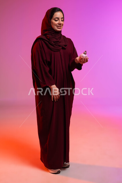 التعطر والتطيب بالعود والزيوت العطرية, التسويق لمنتجات العطور والمستحضرات النسائية, مفهوم الأناقة والاهتمام بالمظهر الخارجي, بورتريه لامرأة عربيه سعودية خليجية محجبة مبتسمة تمسك بيدها زجاجة العطر, صورة لكامل الجسم، خلفية ملونة