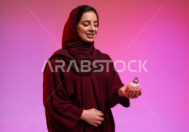 مفهوم الأناقة والاهتمام بالمظهر الخارجي, التعطر والتطيب بالعود والزيوت العطرية, التسويق لمنتجات العطور والمستحضرات النسائية, بورتريه مقرب لامرأة عربيه سعودية خليجية محجبة مبتسمة تمسك بيدها زجاجة العطر، خلفية ملونة