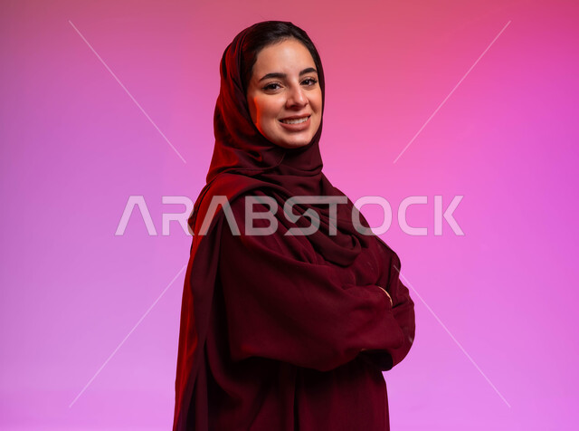 Attention to elegance and external appearance, standing with crossed hands, close-up portrait of a smiling Saudi Gulf Arab woman wearing the abaya and hijab, looking at the camera with gestures of pleasure and happiness, colorful background.