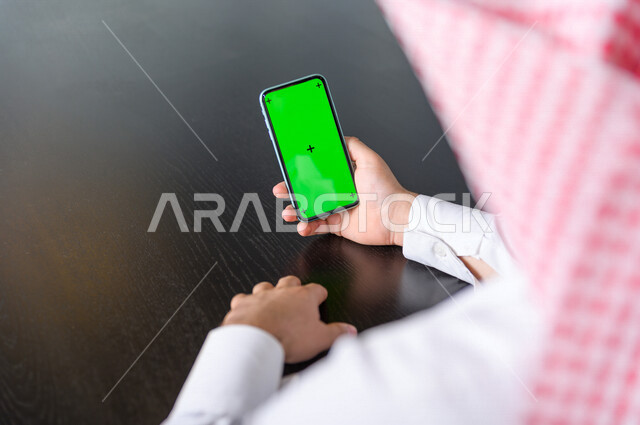 رجل أعمال سعودي خليجي يستخدام هاتفه المحمول بخلفية خضراء كروما ، تقنية و حاسب ، استخدام تقنية السمارت على الهاتف المحمول ليتحكم بالتلفاز