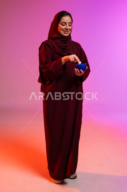الاهتمام بالأناقة والمظهر الخارجي، العناية بنضارة وتحسين البشرة، بورتريه لسيدة عربية خليجية سعودية محجبة مبتسمة ترتدي العباءة تمسك بيدها كريم ترطيب اليدين بإيماءات السرور، استخدام كريمات ومكونات تجميلية، صورة الجسم بالطول الكامل، خلفية ملونة