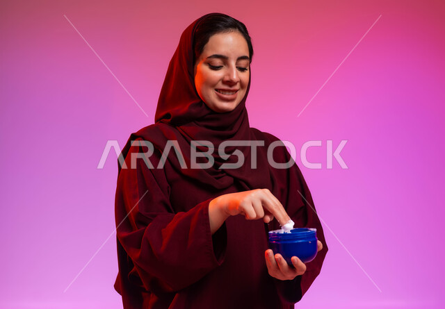 العناية بنضارة وتحسين البشرة، الاهتمام بالأناقة والمظهر الخارجي، بورتريه مقرب لسيدة عربية خليجية سعودية محجبة مبتسمة ترتدي العباءة تمسك بيدها كريم ترطيب اليدين بإيماءات السرور، استخدام كريمات ومكونات تجميلية، خلفية ملونة