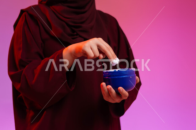 The concept of taking care of the freshness and improvement of the skin, attention to elegance and external appearance, a close-up portrait of a Saudi Arabian Gulf woman wearing an abaya, holding a moisturizing hand cream in her hand, using creams and cosmetic ingredients, a colored background.
