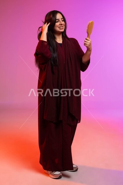 ترتيب وتمشيط الشعر، المحافظة على شعر صحي وأنيق، العناية والاهتمام بالجمال والأناقة والمظهر الخارجي، بورتريه لشابة عربية خليجية سعودية تمسك بيدها فرشاة الشعر ( المشط ) بتعابير وإيماءات تدل على السعادة والسرور، صورة لكامل الجسم، خلفية ملونة