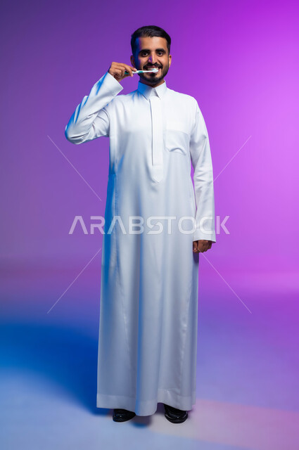 روتين نظافة الفم اليومية، النظر إلى الكاميرا بإيماءات السرور، بورتريه لرجل عربي خليجي سعودي يقوم بتنظيف أسنانه باستخدام الفرشاة والمعجون، مفهوم العناية والاهتمام بصحة ونظافة الأسنان، تجنب التسوس وأمراض اللثة، صورة الجسم بالكامل، خلفية ملونة