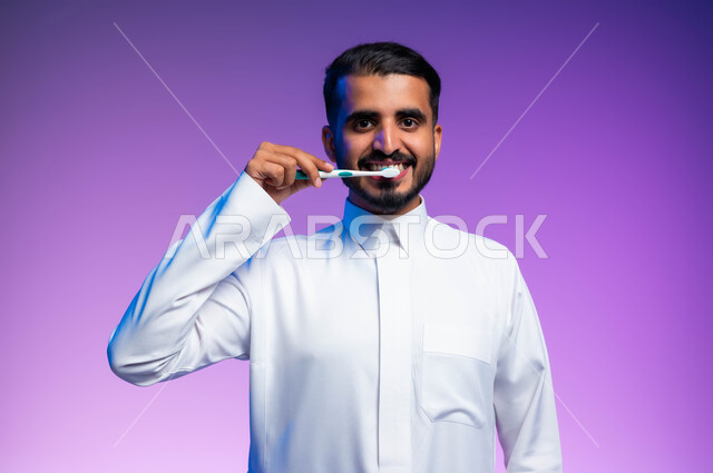 النظر إلى الكاميرا بإيماءات السرور، روتين نظافة الفم اليومية، بورتريه مقرب لرجل عربي خليجي سعودي يقوم بتنظيف أسنانه باستخدام الفرشاة والمعجون، مفهوم العناية والاهتمام بصحة ونظافة الأسنان، تجنب التسوس وأمراض اللثة، خلفية ملونة