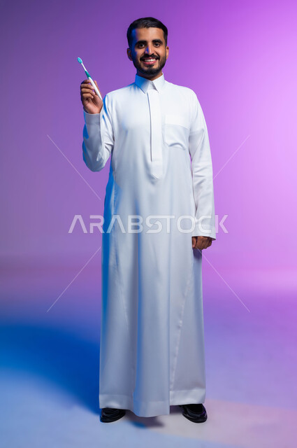 تجنب التسوس وأمراض اللثة، روتين نظافة الفم اليومية، النظر إلى الكاميرا بإيماءات السرور، بورتريه لرجل عربي خليجي سعودي يمسك بيده فرشاة أسنانه، مفهوم العناية والاهتمام بصحة ونظافة الأسنان، صورة الجسم بالكامل، خلفية ملونة