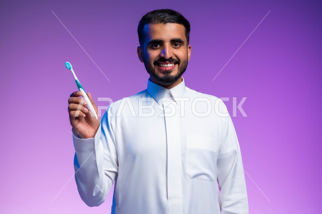 تجنب التسوس وأمراض اللثة، روتين نظافة الفم اليومية، النظر إلى الكاميرا بإيماءات السرور، بورتريه مقرب لرجل عربي خليجي سعودي يمسك بيده فرشاة أسنانه، مفهوم العناية والاهتمام بصحة ونظافة الأسنان، خلفية ملونة