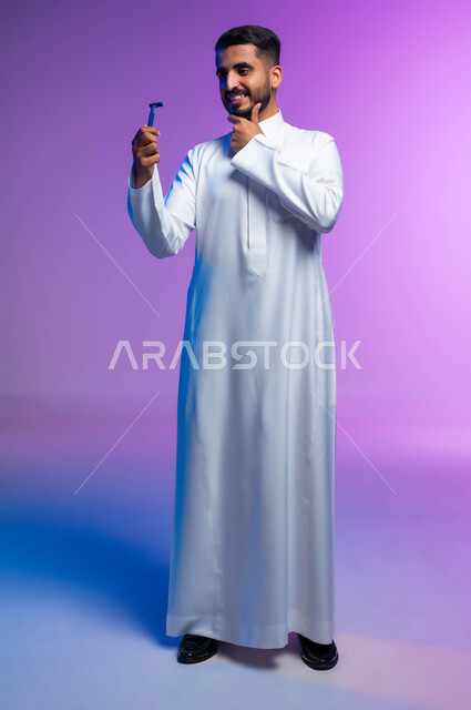 الحفاظ على الأناقة والنظافة الشخصية، العناية بالبشرة والاهتمام بالمظهر الخارجي، بورتريه لرجل عربي خليجي سعودي يرتدي الثوب التقليدي يمسك بيده شفرة الحلاقة الرجالية وينظر إليها بإيماءات الإعجاب والسرور، صورة الجسم بالكامل، خلفية ملونة