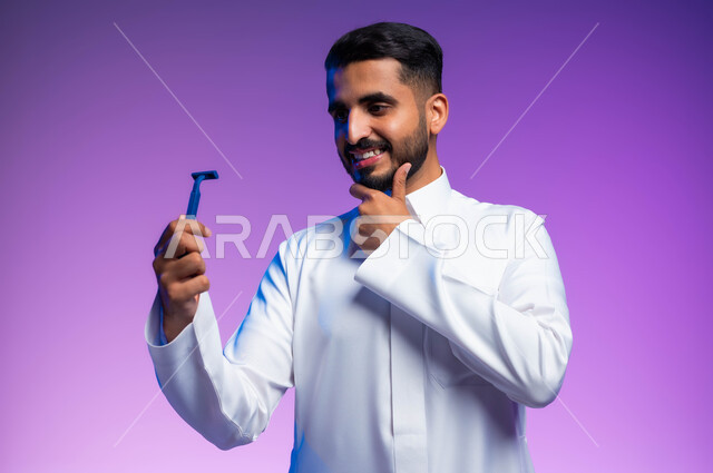 العناية بالبشرة والاهتمام بالمظهر الخارجي، المحافظة على الأناقة والنظافة الشخصية، بورتريه مقرب لرجل عربي خليجي سعودي يرتدي الثوب التقليدي يمسك بيده شفرة الحلاقة الرجالية وينظر إليها بإيماءات الإعجاب والسرور، خلفية ملونة