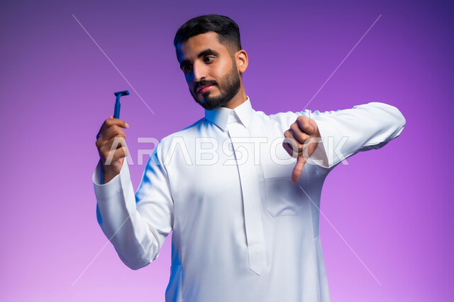الإشارة بعلامة عدم القبول والإعجاب، الحفاظ على الأناقة والنظافة الشخصية، العناية بالبشرة والاهتمام بالمظهر الخارجي، بورتريه مقرب لرجل عربي خليجي سعودي يرتدي الثوب التقليدي يمسك بيده شفرة الحلاقة الرجالية، خلفية ملونة