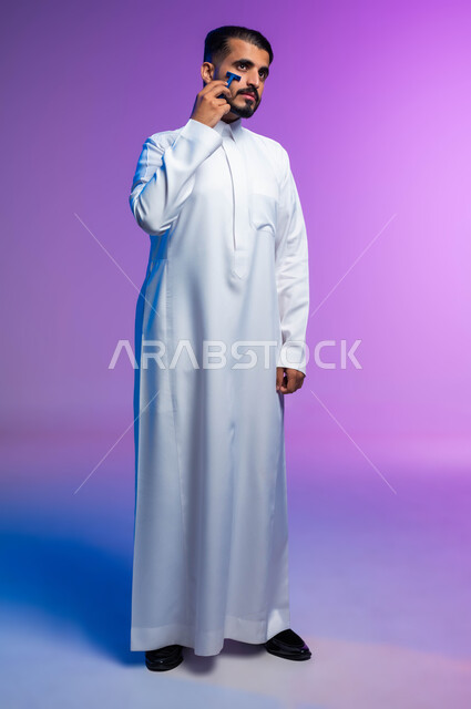 المحافظة على الاناقة والنظافة الشخصية، مفهوم العناية بالبشرة والاهتمام بالمظهر الخارجي، بورتريه لرجل عربي خليجي سعودي يرتدي الثوب التقليدي يقوم بإزالة شعر الوجه باستخدام شفرة الحلاقة الرجالية، صورة لكامل الجسم، خلفية ملونة