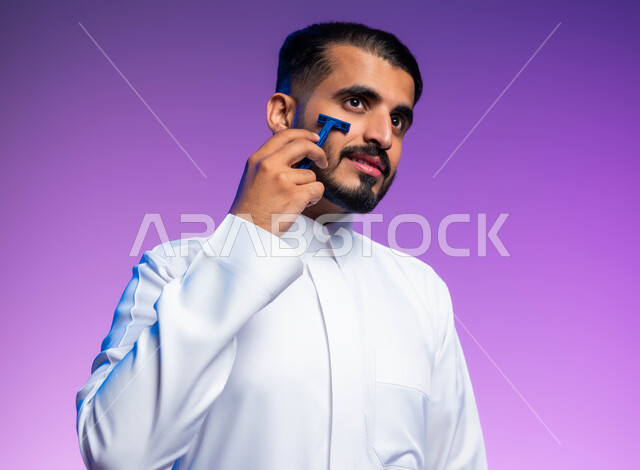 مفهوم العناية بالبشرة والاهتمام بالمظهر الخارجي، المحافظة على الأناقة والنظافة الشخصية، بورتريه مقرب لرجل عربي خليجي سعودي يرتدي الثوب التقليدي يقوم بإزالة شعر الوجه باستخدام شفرة الحلاقة الرجالية بإيماءات التمعن والتركيز، خلفية ملونة