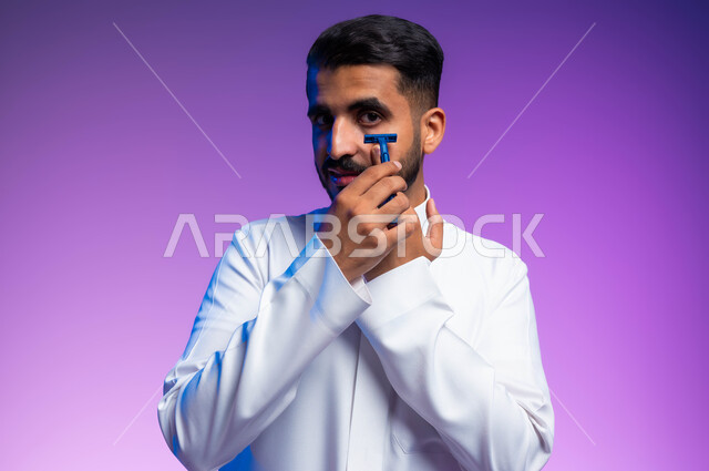مفهوم العناية بالبشرة والاهتمام بالمظهر الخارجي، المحافظة على الأناقة والنظافة الشخصية، بورتريه مقرب لرجل عربي خليجي سعودي يرتدي الثوب التقليدي يقوم بإزالة شعر الوجه باستخدام شفرة الحلاقة الرجالية بإيماءات التمعن والتركيز، خلفية ملونة