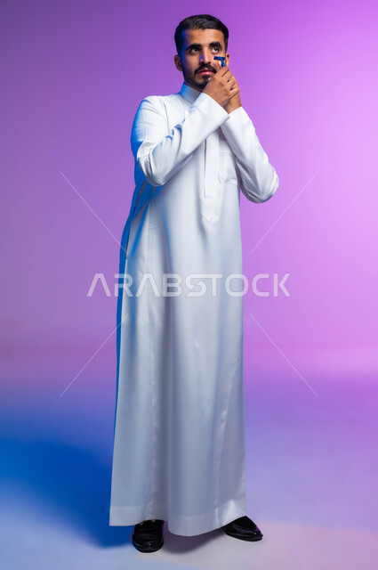 الاهتمام بالمظهر الخارجي، بورتريه لرجل عربي خليجي سعودي يرتدي الثوب التقليدي يقوم بإزالة شعر الوجه باستخدام شفرة الحلاقة الرجالية، مفهوم العناية بالبشرة والنظافة الشخصية، صورة لكامل الجسم، خلفية ملونة