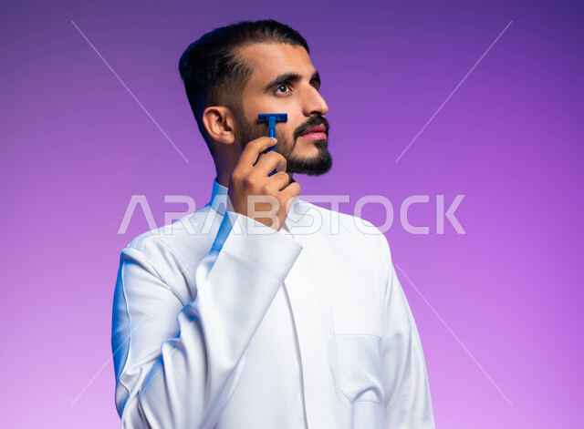 العناية بالبشرة والنظافة الشخصية، الاهتمام بالمظهر الخارجي، بورتريه مقرب لرجل عربي خليجي سعودي يرتدي الثوب التقليدي يقوم بإزالة شعر الوجه باستخدام شفرة الحلاقة الرجالي، خلفية ملونة