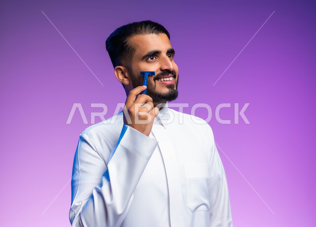 الاهتمام بالمظهر الخارجي، العناية بالبشرة والنظافة الشخصية، بورتريه مقرب لرجل عربي خليجي سعودي يرتدي الثوب التقليدي يقوم بإزالة شعر الوجه باستخدام شفرة الحلاقة الرجالي بإيماءات السرور، خلفية ملونة