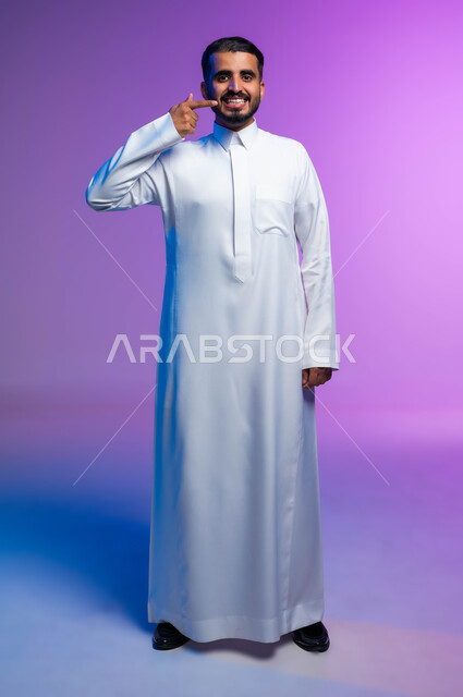 العناية والاهتمام بالصحة الفموية، إبتسامة هوليود باستخدام قشور فينير من الخزف، بورتريه لرجل عربي خليجي سعودي مبتسم يرتدي الثوب التقليدي ينظر إلى الكاميرا ويشير بإصبعه السبابة على أسنانه، صورة الجسم بالطول الكامل, خلفية ملونة.