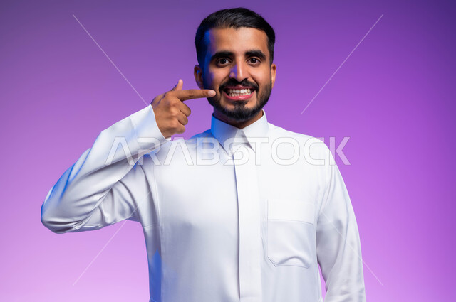 إبتسامة هوليود باستخدام قشور فينير من الخزف، بورتريه مقرب لرجل عربي خليجي سعودي مبتسم يرتدي الثوب التقليدي ينظر إلى الكاميرا ويشير بإصبعه السبابة على أسنانه، العناية والاهتمام بالصحة الفموية، خلفية ملونة.