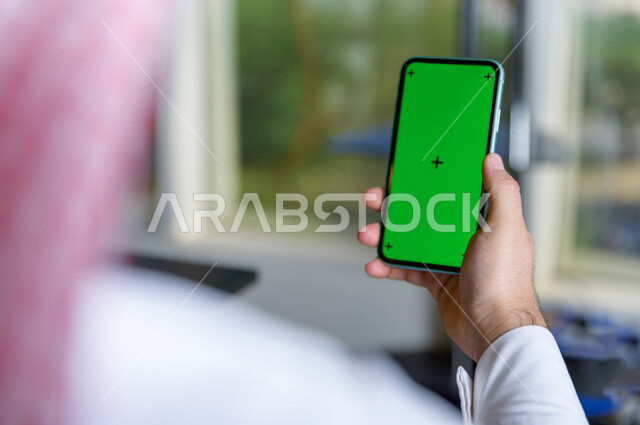 رجل أعمال سعودي خليجي يستخدام هاتفه المحمول بخلفية خضراء كروما ، تقنية و حاسب ، استخدام تقنية السمارت على الهاتف المحمول ليتحكم بالتلفاز