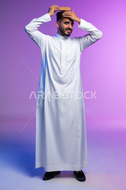 Looking at the camera with expressions and gestures that indicate confidence and pleasure, arranging and combing hair, the concept of care and attention to beauty, elegance and external appearance, a portrait of a Saudi Arabian Gulf man styling his hair using a hairbrush (comb), full-body portrait, colored background.