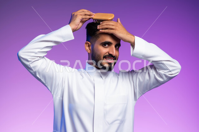 النظر إلى الكاميرا بتعابير وإيماءات تدل على الثقة والسرور، ترتيب وتمشيط الشعر، مفهوم العناية والاهتمام بالجمال والأناقة والمظهر الخارجي، بورتريه مقرب لرجل عربي خليجي سعودي يقوم بتصفيف شعره باستخدام فرشاة الشعر ( المشط )، خلفية ملونة