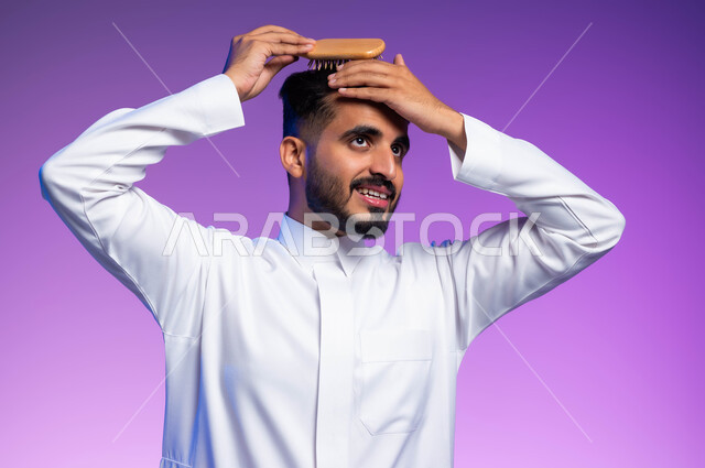 مفهوم العناية والاهتمام بالجمال والأناقة والمظهر الخارجي، بورتريه مقرب لرجل عربي خليجي سعودي يقوم بتصفيف شعره باستخدام فرشاة الشعر ( المشط )، تعابير وإيماءات تدل على السعادة والسرور، خلفية ملونة