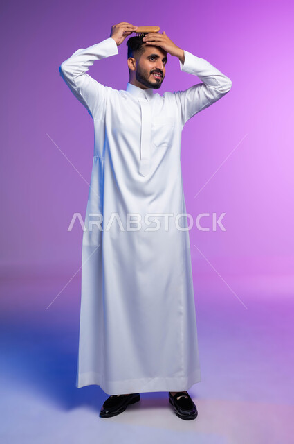 تمشيط وترتيب الشعر، مفهوم العناية والاهتمام بالجمال والأناقة والمظهر الخارجي، بورتريه لرجل عربي خليجي سعودي يقوم بتصفيف شعره باستخدام فرشاة الشعر ( المشط ) بتعابير وإيماءات تدل على السعادة والسرور، صورة الجسم بالكامل، خلفية ملونة