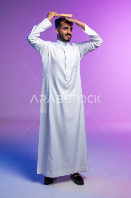 تمشيط وترتيب الشعر، مفهوم العناية والاهتمام بالجمال والأناقة والمظهر الخارجي، بورتريه لرجل عربي خليجي سعودي يقوم بتصفيف شعره باستخدام فرشاة الشعر ( المشط ) بتعابير وإيماءات تدل على السعادة والسرور، صورة الجسم بالكامل، خلفية ملونة
