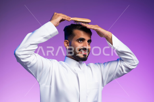 مفهوم العناية والاهتمام بالجمال والأناقة والمظهر الخارجي، بورتريه مقرب لرجل عربي خليجي سعودي يقوم بتصفيف شعره باستخدام فرشاة الشعر ( المشط )، تعابير وإيماءات تدل على السعادة والسرور، خلفية ملونة