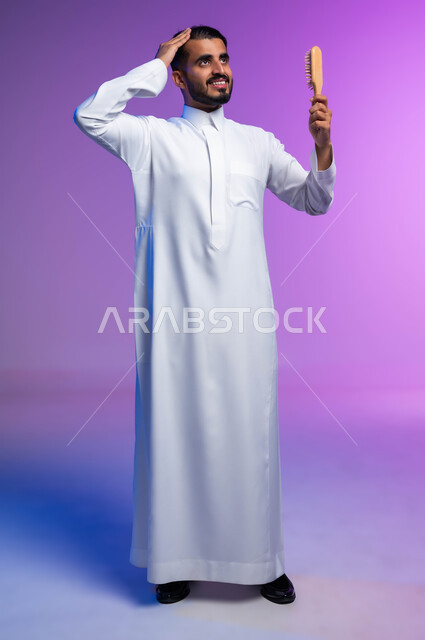 ترتيب وتمشيط الشعر، مفهوم العناية والاهتمام بالجمال والأناقة والمظهر الخارجي، بورتريه لرجل عربي خليجي سعودي يقوم بتصفيف شعره باستخدام فرشاة الشعر ( المشط ) بتعابير وإيماءات تدل على السعادة والسرور، صورة الجسم بالكامل، خلفية ملونة