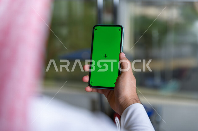 رجل أعمال سعودي خليجي يستخدام هاتفه المحمول بخلفية خضراء كروما ، تقنية و حاسب ، استخدام تقنية السمارت على الهاتف المحمول ليتحكم بالتلفاز