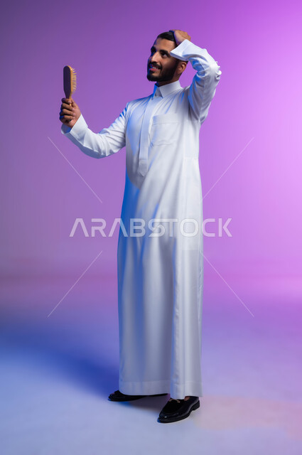 تمشيط وترتيب الشعر، مفهوم العناية والاهتمام بالجمال والأناقة والمظهر الخارجي، بورتريه لرجل عربي خليجي سعودي يقوم بتصفيف شعره باستخدام فرشاة الشعر ( المشط ) بتعابير وإيماءات تدل على السعادة والسرور، صورة الجسم بالكامل، خلفية ملونة