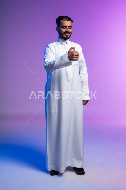 عطورات خليجية سعودية, مفهوم الاناقة والاهتمام بالمظهر الخارجي, استخدام الزيوت العطرية الطبيعية, بورتريه لرجل عربي خليجي سعودي يرتدي الثوب التقليدي يقوم بوضع العطر, التسويق لمنتجات العطور, صورة الجسم بالكامل, خلفية ملونة