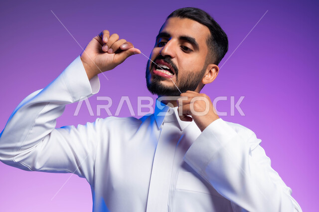 المحافظة على ابتسامة بيضاء، الاهتمام بالأناقة والمظهر الخارجي، بورتريه مقرب لرجل عربي خليجي سعودي يقوم بتنظيف أسنانه باستخدام الخيط السني، مفهوم العناية بصحة الفم والأسنان، خلفية ملونة