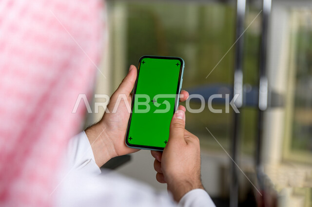 رجل أعمال سعودي خليجي يستخدام هاتفه المحمول بخلفية خضراء كروما ، تقنية و حاسب ، استخدام تقنية السمارت على الهاتف المحمول ليتحكم بالتلفاز