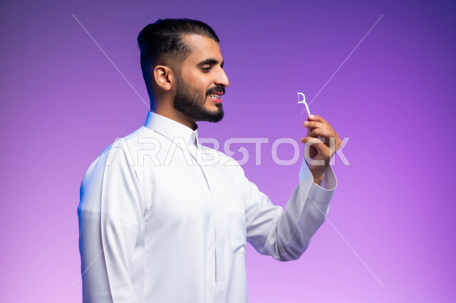 العناية بالصحة الفموية، التأكد من نظافة الأسنان بعد تناول الطعام، الاهتمام بالأناقة والمظهر الخارجي، المحافظة على ابتسامة بيضاء، بورتريه مقرب من الجانب لرجل عربي خليجي سعودي يقوم بتنظيف أسنانه باستخدام الخيط السني، خلفية ملونة
