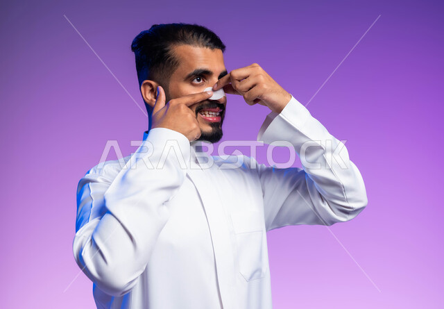 استخدام ماسكات تنظيف المسام ومكونات تجميلية، بورتريه مقرب لرجل عربي خليجي سعودي يرتدي الثوب التقليدي يضع لصقات الأنف للتخلص من الرؤوس السوداء والجلد الميت، مفهوم العناية والاهتمام بنضارة وتحسين البشرة، خلفية ملونة