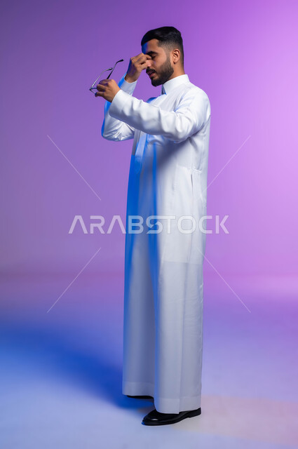 الاناقة والاهتمام بالمظهر الخارجي، بورتريه من الجانب لرجل عربي خليجي سعودي يرتدي الثوب التقليدي يمسك النظاره الطبيه يضع يده علىوجهه بإيماءاتالتعب والإرهاق، ارتداء النظارات الخاصة بضعف النظر، مفهوم تعديل وتحسين الرؤية، صورة لكامل الجسم، خلفية ملونة